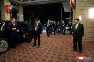 a man in a dark suit steps out of a black sedan as staff and security line up outside a building with north korean flags overhead